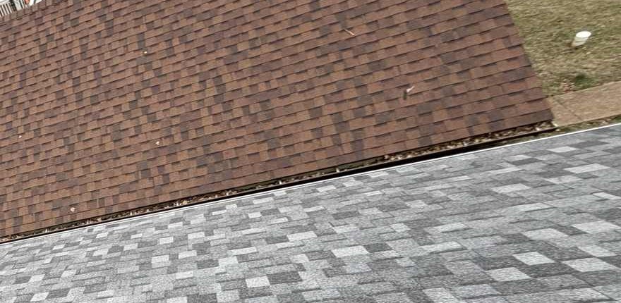Looking down at the junction between two roof planes: an old weathered brown shingle roof above and a newly laid grey and charcoal architectural shingle roof below, with the open gutter running diagonally between them holding a continuous line of dark leaf litter and twigs along its whole visible length