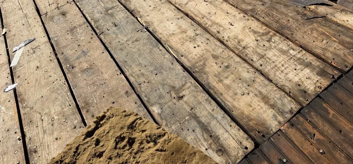 A roof plane stripped back to its original wide wooden plank decking, the boards weathered grey and separated by open gaps, studded with hundreds of old nail shanks, with a heaped pile of loose shingle granules in the centre foreground and two pale metal offcuts at the left edge