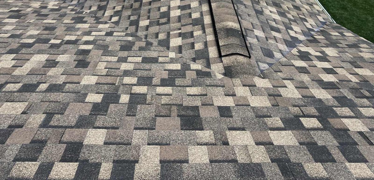 A brown, tan and charcoal multi tone architectural shingle roof filling the frame, with a hip running up to a capped ridge in the upper right where ridge cap shingles sit over a dark ridge vent strip, and the two roof planes falling away either side