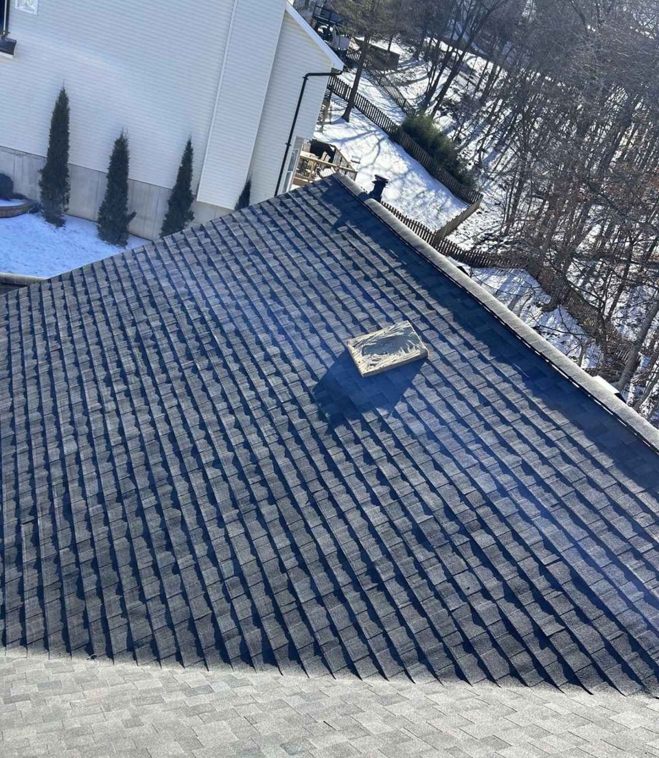 A large hip roof plane of grey architectural shingles photographed in low winter sun, so every course throws a hard shadow line down the slope, with a small metal vent near the ridge and a torn shingle wrapper and an offcut lying loose on the surface. A second, flatter lit plane of the same shingle meets it across the bottom of the frame. Beyond the ridge are a cream lap sided wall on a stone base, three clipped upright conifers, a wooden deck and rail, a picket fence, lying snow and bare woodland